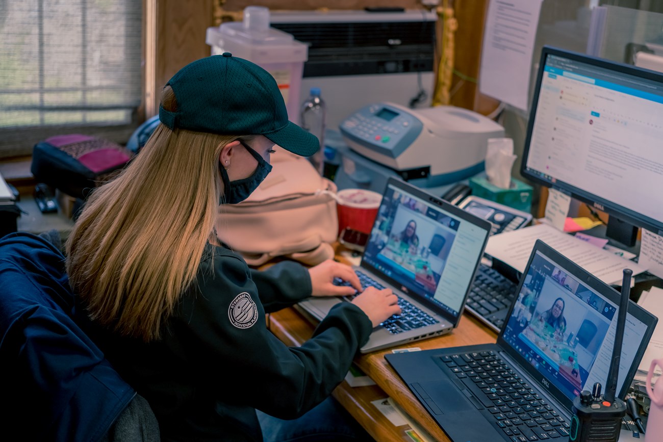 A woman in a park service green sweater & ballcap sits before two laptops & a computer screen