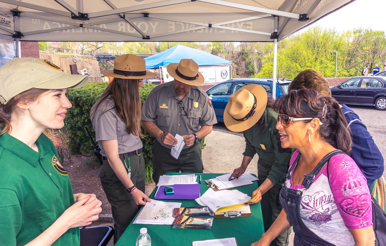 Uniformed park rangers in flat hats & a volunteer in green assist several people with information at a table stocked with brochures, maps, & sign-up clipboards under a tent
