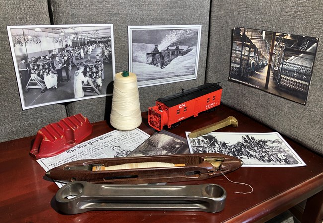 A corner table & the walls behind it hold historic photos, newspaper clippings, & artifacts highlighting Paterson's railroad, silk, & aircraft industries, such as a loom shuttle, spool of thread, engine parts, a rotary snowplow model, & a golden spike.