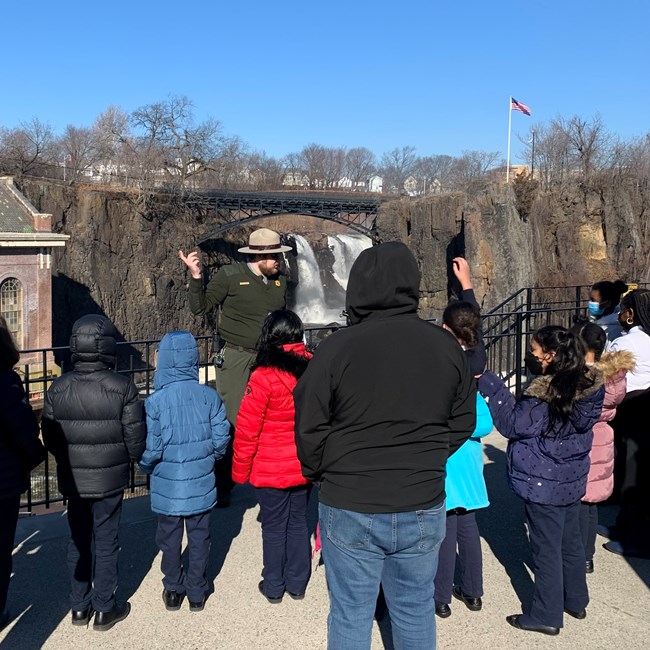 A bearded park ranger in winter felt flat hat and green sweater leading a tour of a group of children in coats in front of a 77 ft. waterfall falling between dark basalt cliffs & below an arched black metal bridge