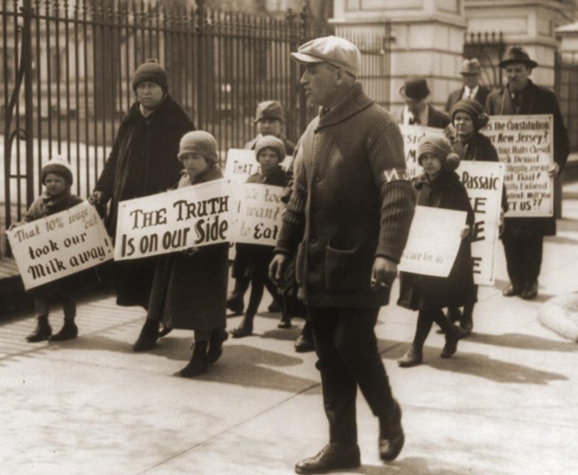 1913 photo of men, a woman, & numerous children marching with strike picket signs