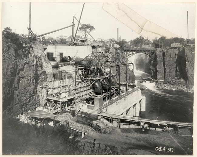 Black & white photo dated Oct. 4th 1913 shows construction of a hydroelectric plant beside a 77 ft. waterfall framed by dark basalt cliffs & and arched metal bridge. The building's metal frame houses massive metal pipes running into a cut in the cliff