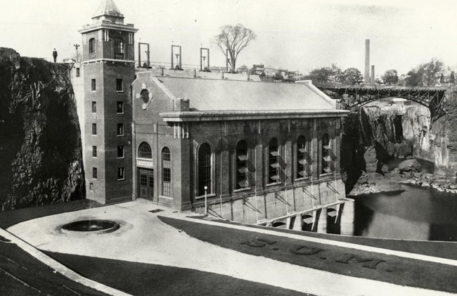 Black & white photo of a brick hudroelectric plant w/ peaked roof & stair tower built into a cliff beside a dry 77 ft. waterfall framed by a black arched metal bridge. Landscaping beside it spells "SUM" in shrubs, & a fountain bubbles before it