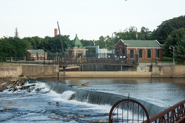 The Passaic River flows right to left over a dam. The intake to a brick hydroelectric power plant, spanned by a short arched metal bridge, sits adjacent.