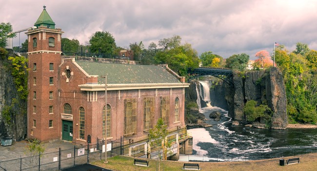 A brick hydroelectric power plant with tall stair tower & green sloped roof stands beside a 77 ft. waterfall framed by dark basalt cliffs & an arched black metal bridge