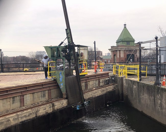 Workers use a large, angled green electromechanical arm operated from a crane-like cab to pull debris off of the concrete & wood intake of a hydroelectric plant