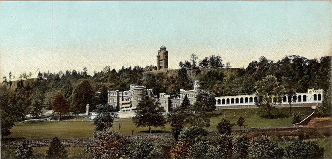 Early 20th Century colorized postcard of a long three story castle with a nearby observation tower on the side of a mountain