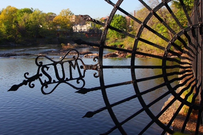 A half-moon black ornamental fence facing left with the letters "S.U.M." in ornate filigree. Facing left, it frames a view of the Passaic River & the upper edge of the Great Falls surrounded by trees