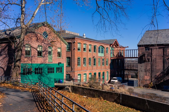 Three story brick mill buildings w/ angled roofs & green windows & doors stand along a dry water channel, autumn leaves gathered on the ground