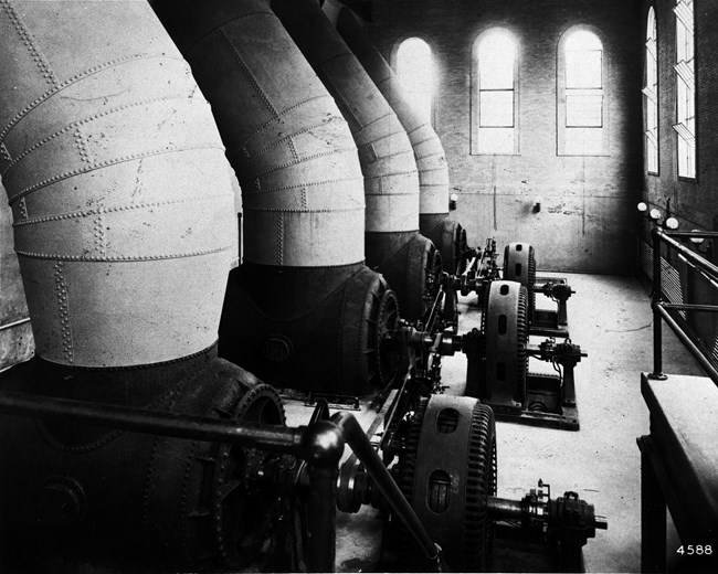 Black & white photo of 4 large metal tubes angled from upper left of a tall building interior into round horizontal turbines connected to disk-like generators, lined up in a row
