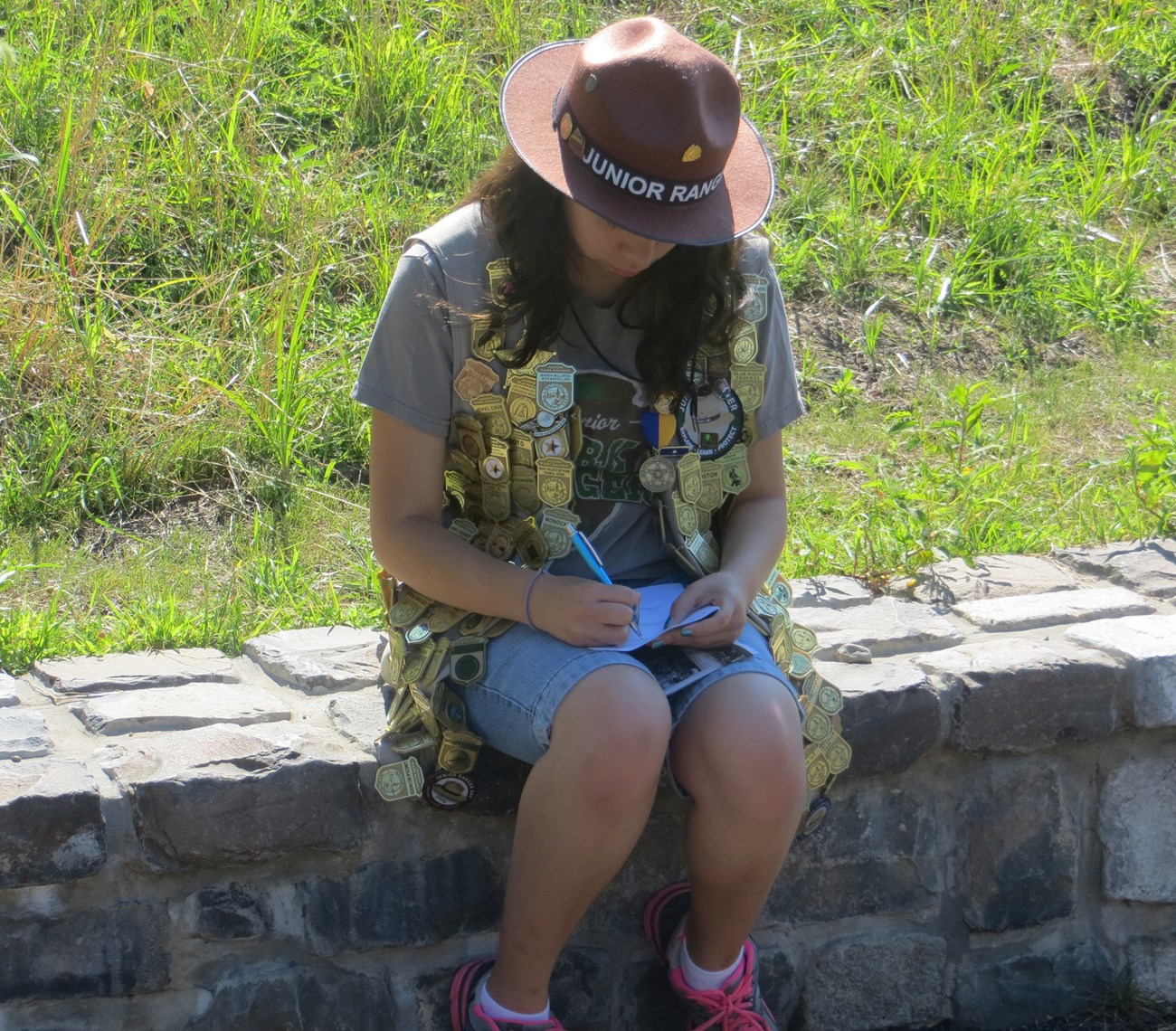 A young girl with a Jr. Ranger flat hat and a vest covered in gold Jr. Ranger badges, patches, & ribbons sits on a wall completing an activity book