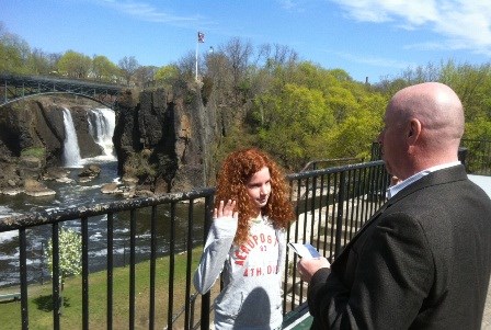 A young red-haired girl standing before the 77 ft. Great Falls where it plunges below an arched metal bridge into the gorge of the Passaic River. Holding her right hand up, she is being sworn in as a Jr. Ranger by a man in a suit.