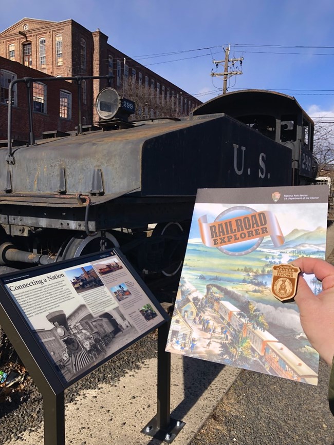 A colorful Railroad Explorer activity book is held up with a wooden badge beside the rear of a black steam locomotive & interpretive wayside sign, set before brick factory buildings
