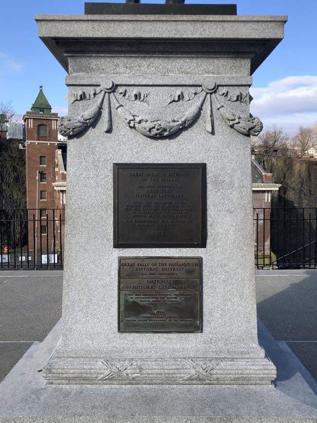 A pair of plaques denoting the establishment of Paterson Great Falls as a National Natural Landmark & Historic District National Historic Landmark in 1967 & 1976, respectively, mounted on a grey statue pedestal