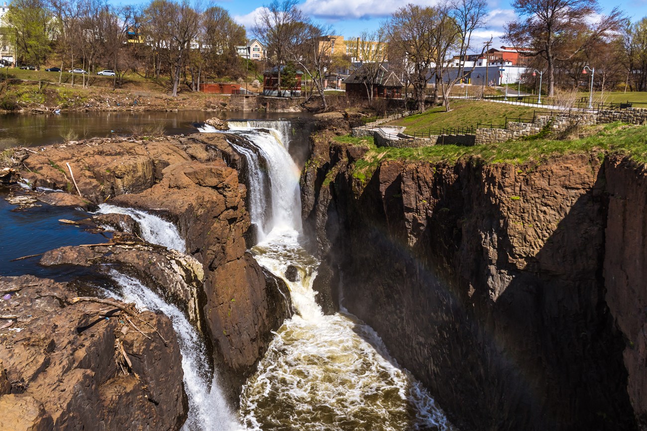 A 77 ft. waterfall plunges into a dark basaltic chasm, a rainbow rising from the spray. At right an overlook & grassy park stretches froward from old brick & concrete buildings in the background