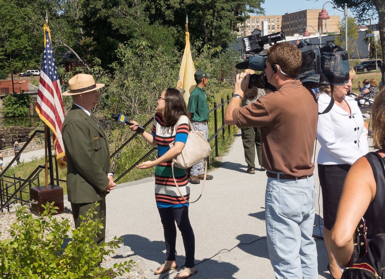 A park ranger in green dress uniform is interviewed by a news crew with film equipment in front of a waterfall overlook flanked by American and State of NJ flags
