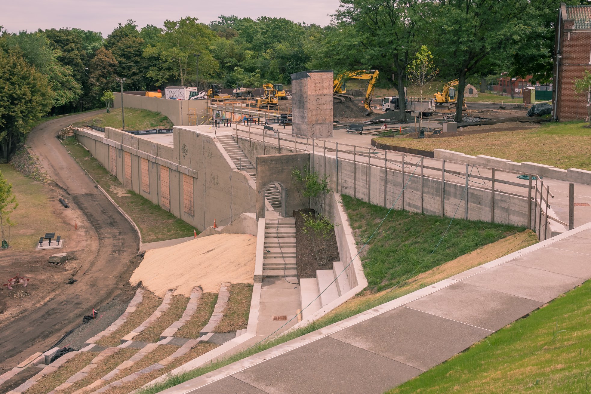 This picture shows the new amphitheater in Overlook Park from above. The arena has multiple stone levels with new green grass growing among them. Yellow construction vehicles can be seen in the background parking lot. Overlook Park Construction Project