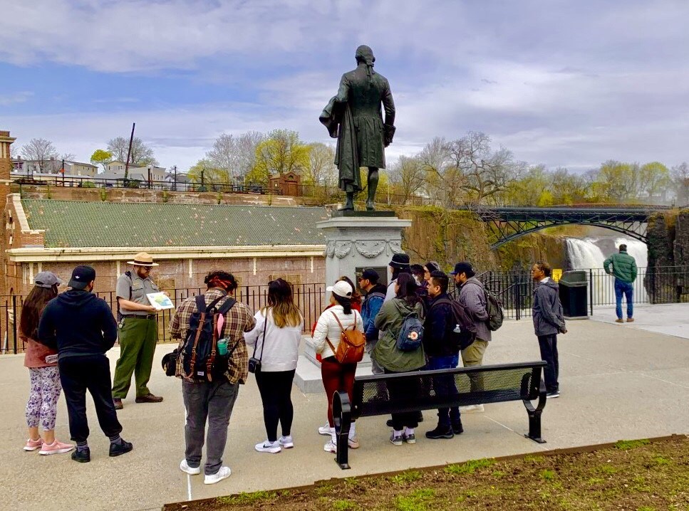 A park ranger in grey & green uniform with straw flat hat shows a historical image to a group of people on a tour standing around a statue of Alexander Hamilton, overlooking the Great Falls, an arched bridge above it & brick hydroelectric plant beside it.