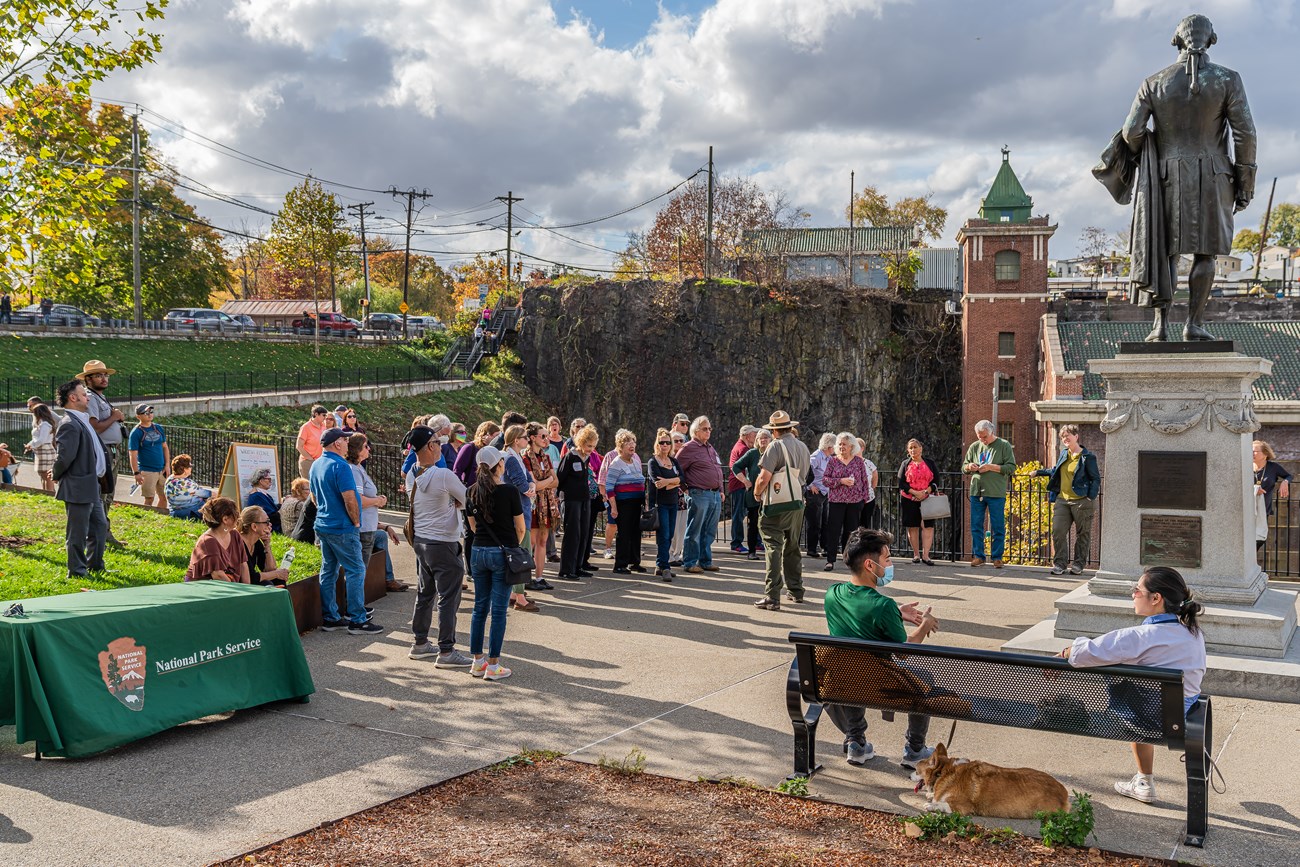 A park ranger in grey & green uniform & straw flat hat speaks to a group of people standing by a statue of Alexander Hamilton, a dark cliff & brick hydroelectric plant in the background. Another ranger stands near a table & other visitors relax with a dog