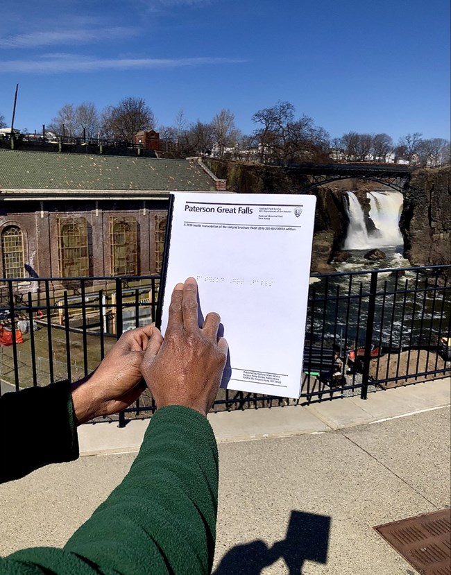 A braille spiral-bound booklet is held before a fence overlooking a waterfall plunging 77 ft. between dark basalt cliffs, framed by a black metal bridge. A brick hydropower plant is visible at left