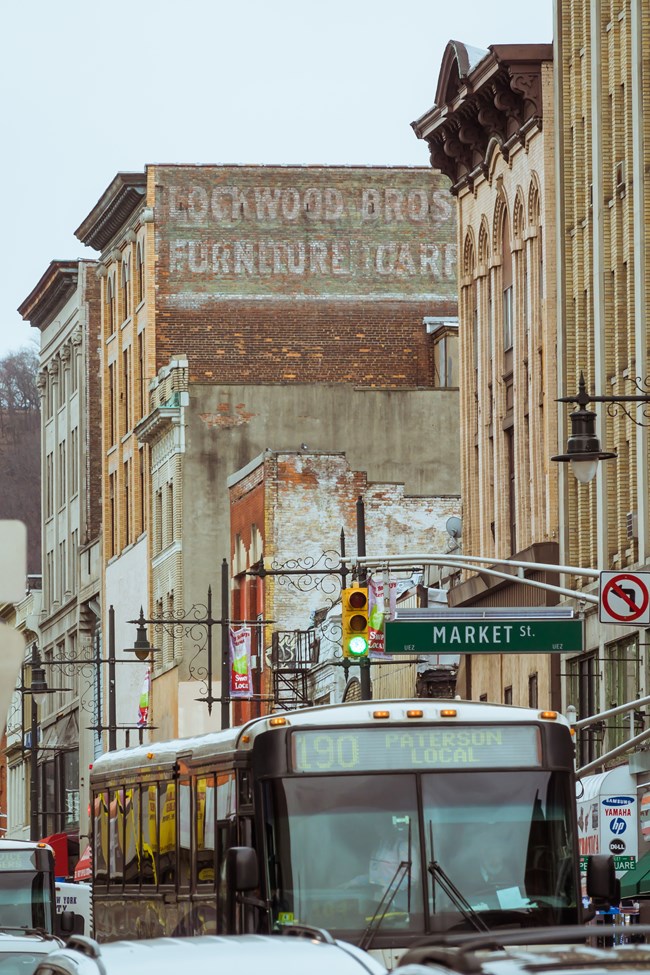 A bus in traffic labeled "190 Paterson Local" passes under a traffic light marked "Market St." - ornate black streetlights line a dense urban core of tall old buildings in red, white, & fawn brick. One has a faded ad for "Lockwood Bros Furniture"