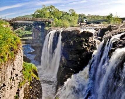 Water plunges down a 77 ft. chasm between dark basalt cliffs, a set of arched metal bridges crossing in the background. Green vegetation frames the left side and background