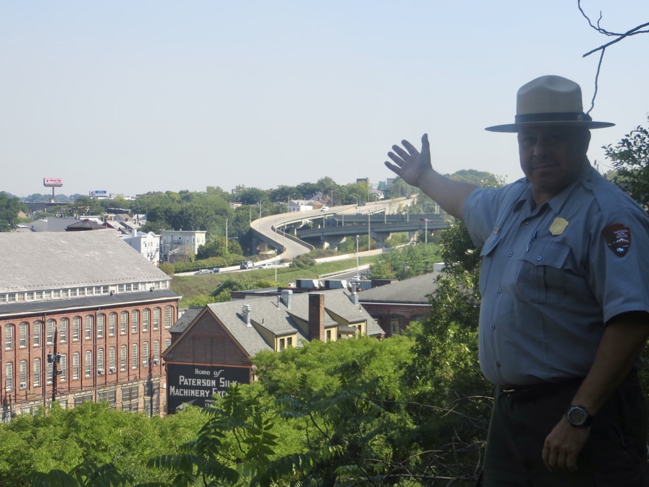 A uniformed Park Ranger gesturing towards historic red brick mill buildings, a highway interchange visible in the background