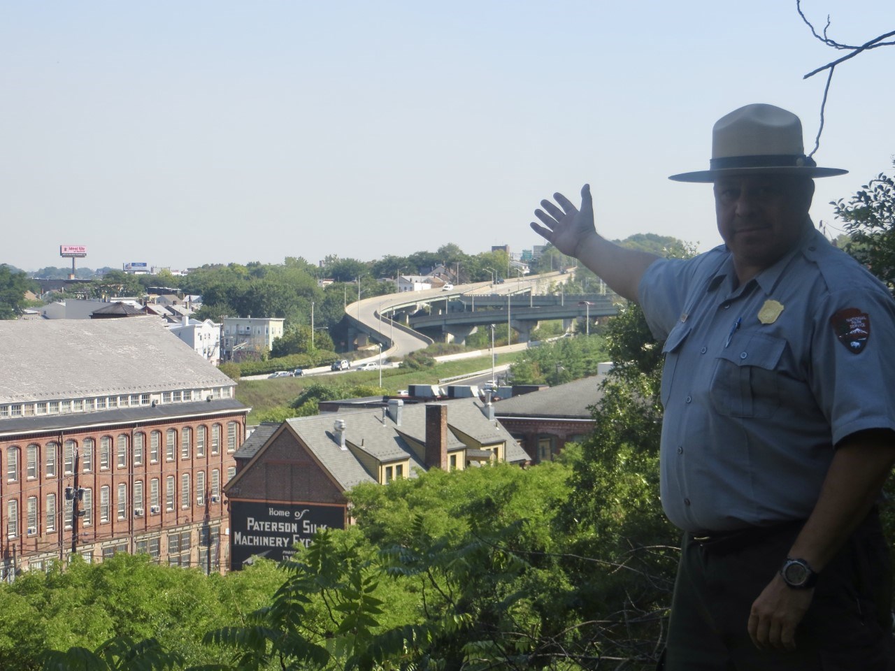 A uniformed Park Ranger gesturing towards historic red brick mill buildings, a highway interchange visible in the background