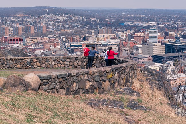 People stand at a stone wall at a mountaintop overlook, the dense urban city of Paterson NJ filling the background below