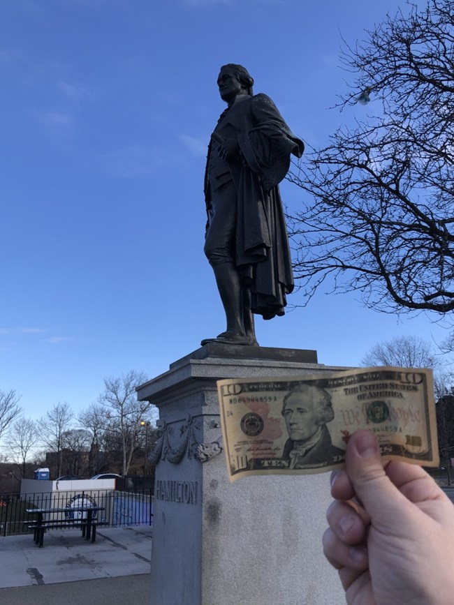 A hand holding a $10 bill up to a statue of Alexander Hamilton, matching the profile image on the money to the statue