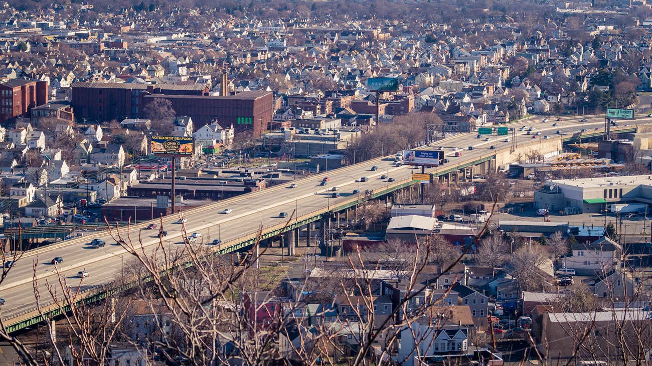 An elevated, 8-lane section of I-80 highway running diagonally from bottom left to top right as it cuts through the dense urban landscape of Paterson NJ