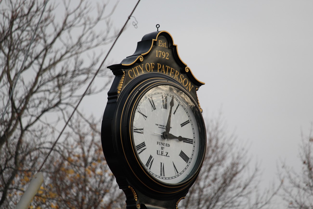 A large black clock with gold lining and scrollwork registering 3:00 set against a greying afternoon sky and winter trees. Text on the frame and face reads "Est. 1792 City of Paterson" & "Funded by U.E.Z."