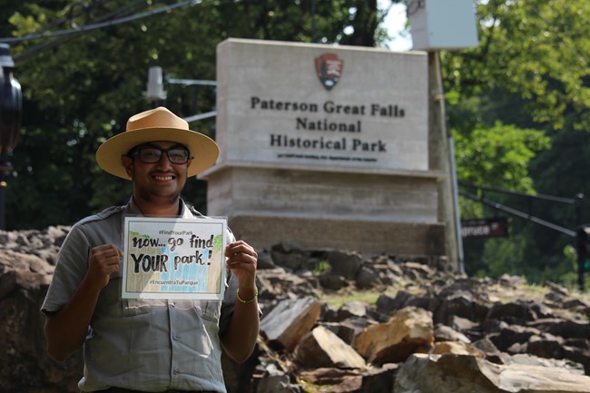 A smiling park ranger with glasses stands before a square stone sign for Paterson Great Falls National Historical Park - he holds a paper sign reading "Now Go Find Your Park! #FindYourPark #EncuentraTuParque" with a drawing of a waterfall