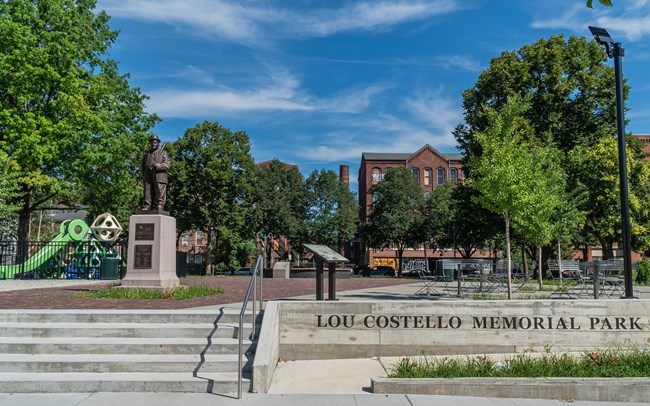 A bronze statue of Lou Costello & a brightly colored set of playground equipment stands at left of an open, tree-shaded courtyard with "Lou Costello Memorial Park" in metal letters on a staircase wall. Brick mill buildings & a smokestack stand at rear