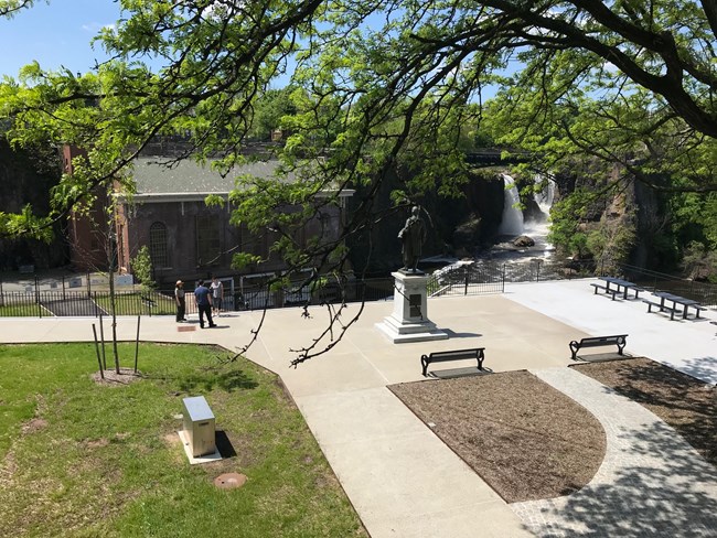 Looking down on the pathways at Overlook Park, the Alexander Hamilton Statue, benches, picnic tables, & falls are visible with a park ranger, & two visitors in the background.  Leafy tree branch at top of picture.
