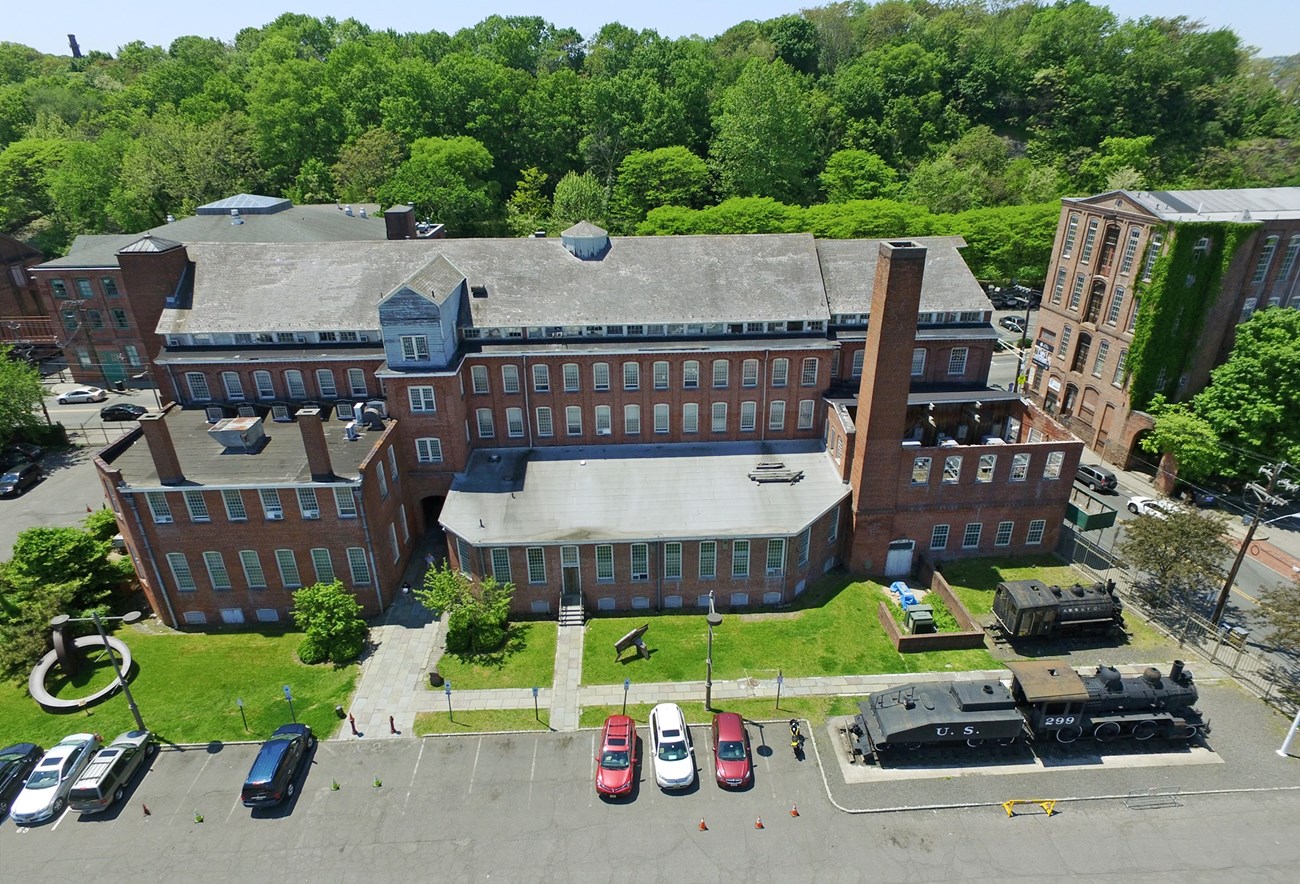 Aerial view of the tall brick 19th century Roger's Locomotive Building with its tall chimney, now the Paterson Museum. Cars in the foreground parking lot sit at right of two steam locomotives