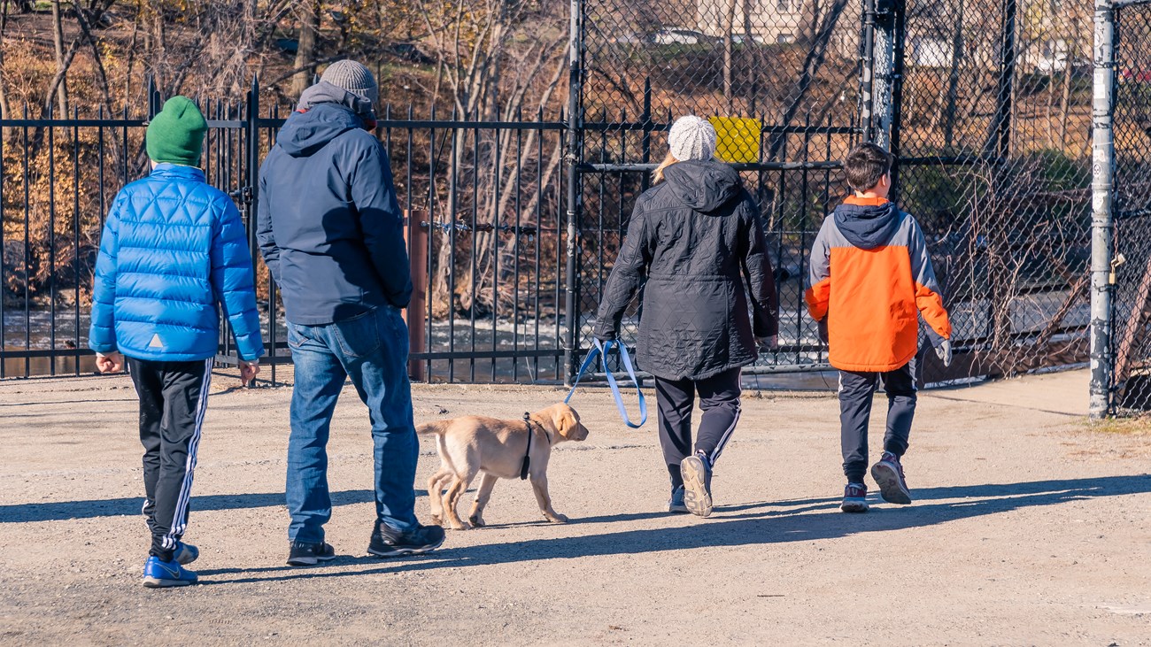 Four people in winter jackets walking a young light-brown dog on a leash, overlooking a river beyond a black metal fence