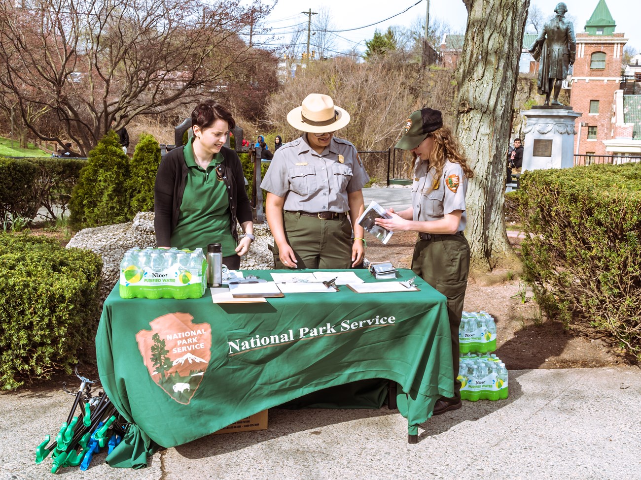 Two park rangers & a green-clad volunteer stand at an info table w/ a green National Park Service covered in flyers, sign-in sheets, & bottled water. One reviews a flyer. Behind them are trees & shrubs, a statue of Alexander Hamilton, & a brick building