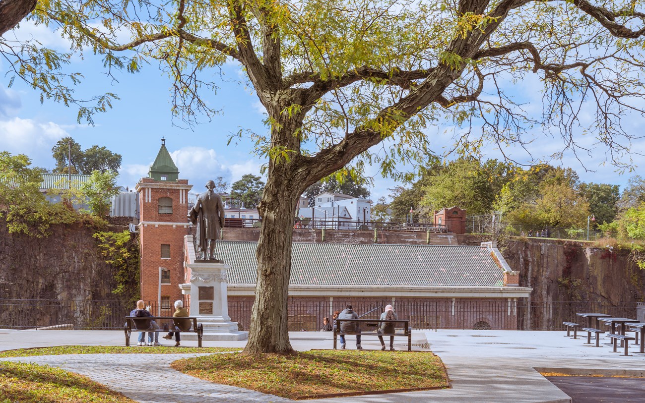Visitors sit at benches shaded by a large tree - they & a statue of Alexander Hamilton face a brick hydroelectric powerplant with a stair tower topped by a green peaked roof along a dark cliffside