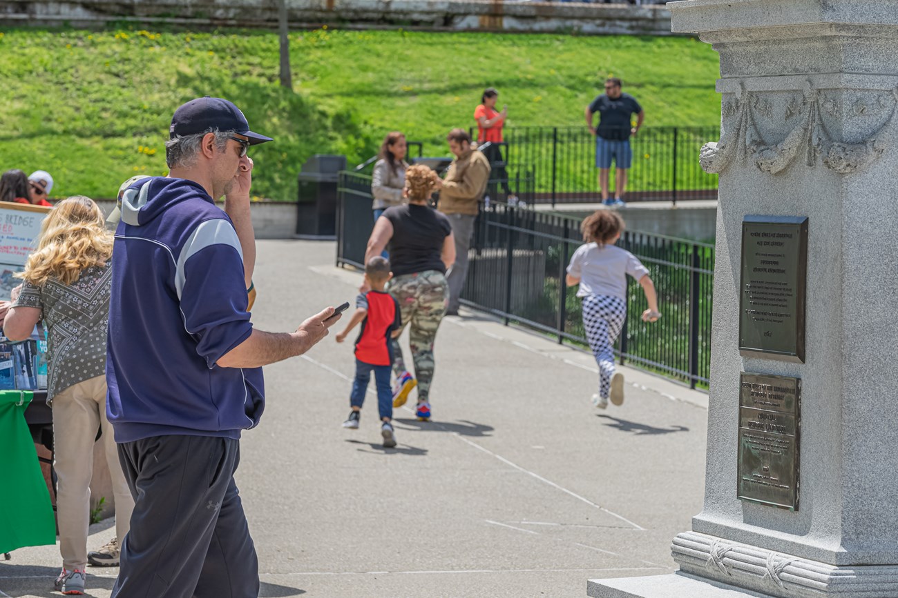 A man checks his phone as he walks past the base of a statue with plaques - other people walk around as a woman checks an information table at left