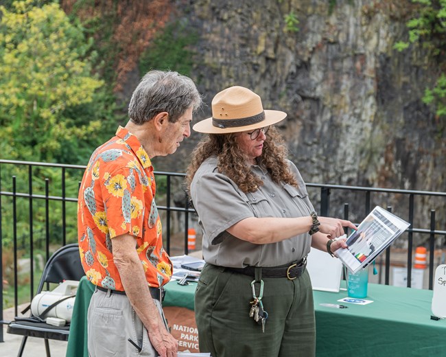 A park ranger in flat hat & uniform points to a laminated information diagram in her hand, providing information for a man in an orange & yellow floral shirt
