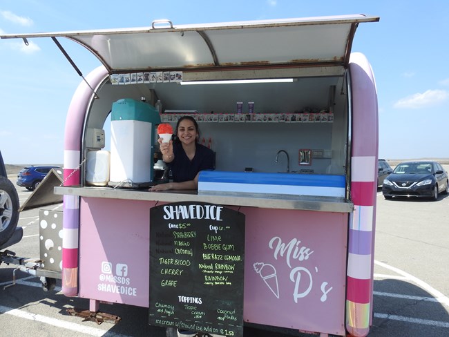 A woman holding out a red snow cone in a pink shaved ice food truck.