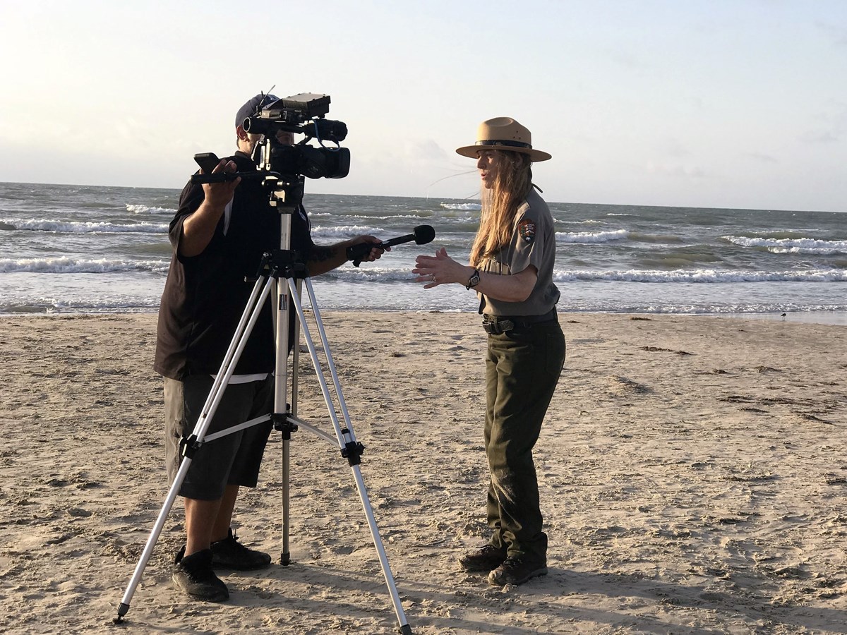 A man interviews and films a National Park Ranger while they stand on the beach with the ocean in the background.
