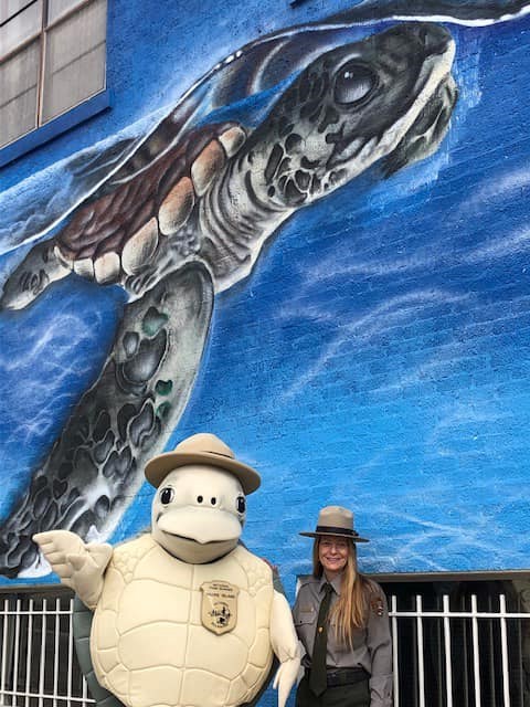 Dr. Shaver and turtle mascot "Seashore Sandy" stand in front of a sea turtle mural painted on the side of a building in downtown Corpus Christi, Texas