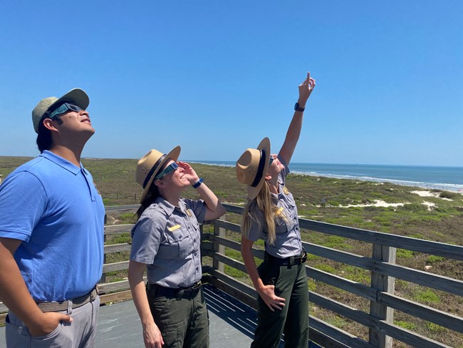 Three people wearing eclipse glasses and looking up at the sun.