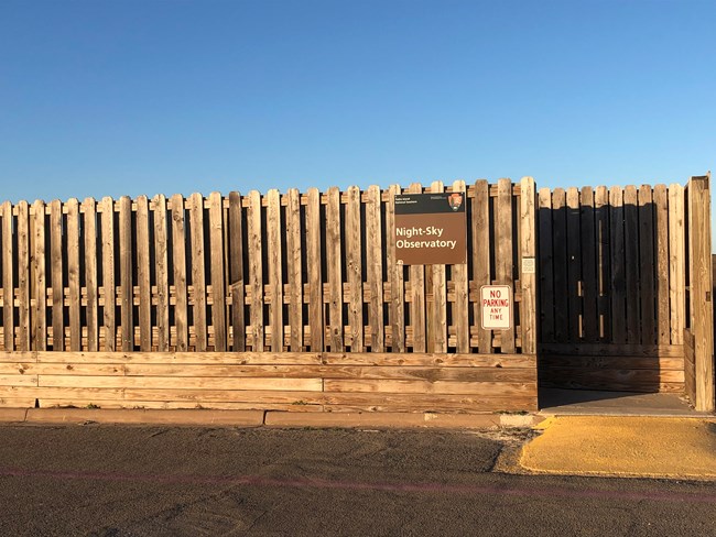 Under a blue sky is one side of an eight-foot-tall wood fence-like structure with a wheelchair ramp and a brown Night Sky Observatory sign