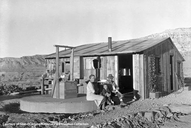 Black and white photo of a family sitting in front of a wooden homestead cabin.