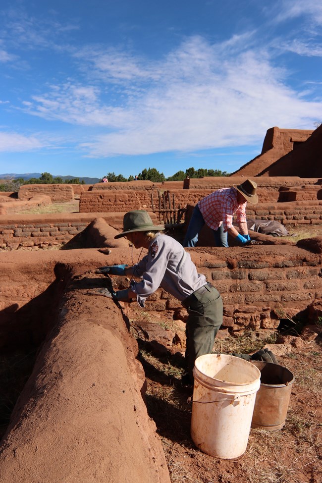 A kneeling woman wearing the National Park Service Uniform daubs mud onto a red adobe wall.
