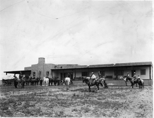 A building with people on horses and wagons gathered in the foreground.