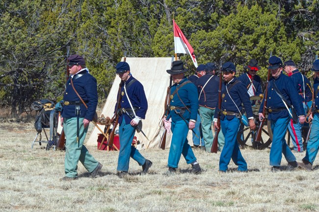 People walking in a group with a firearms around a campsite with trees in the background.